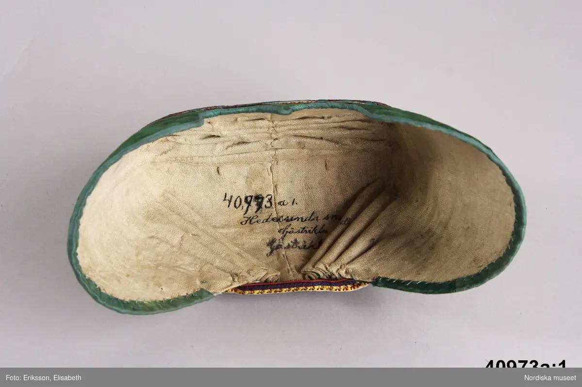 Huvudliggaren: Dräkten "begagnades till början av 1840-talet, linnet av senare årtal."
a.1-2 bindmössa m. stycke
b. tröja längd 35 cm, ärml. 46 cm
c. överdel
d. förkläde
e. kjol
f. 1-2, skor
g. 1-2, bindmössa m. stycke

a.1 BINDMÖSSA av grön sidenatlas med broderi i tambursöm med silke i gult, vitt, rosa, rost, svart och ljusgrönt. Kantad med blågrönt sidenband. Mittsöm över hjässan samt materialskarvar framtill vid sidorna. I nacken 4 och 4 mot mitten lagda veck. Foder av ljus linnelärft. Mönstrat sidenband runt kullen i rödbrunt, lila, gult och vitt.
/Berit Eldvik 2005-09-08