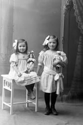 Studioportrett av to jenter med dukker i armene.
