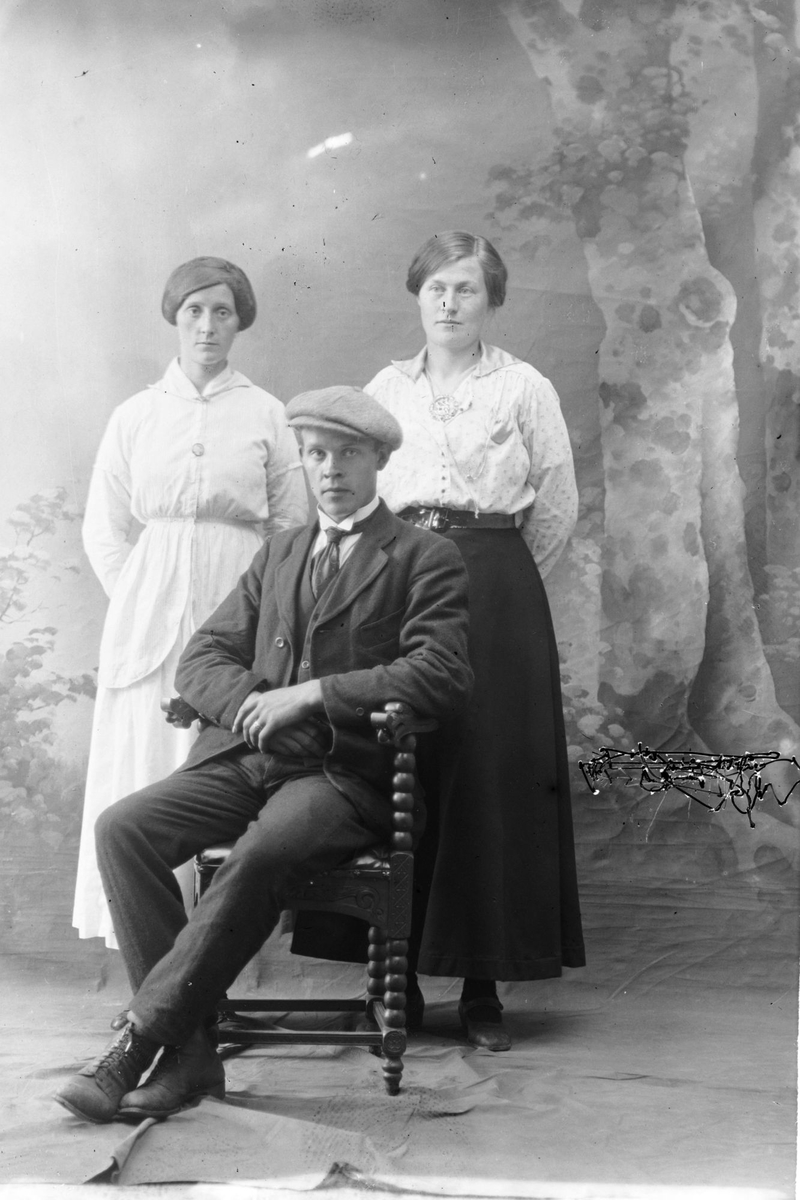 Studioportrett av to kvinner stående bak en mann som sitter.
