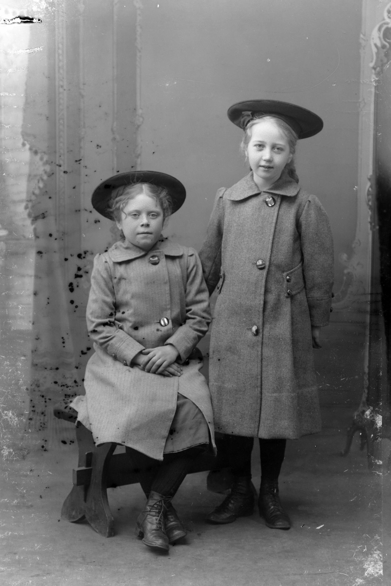 Studioportrett av to unge jenter med store hatter på hodet.
