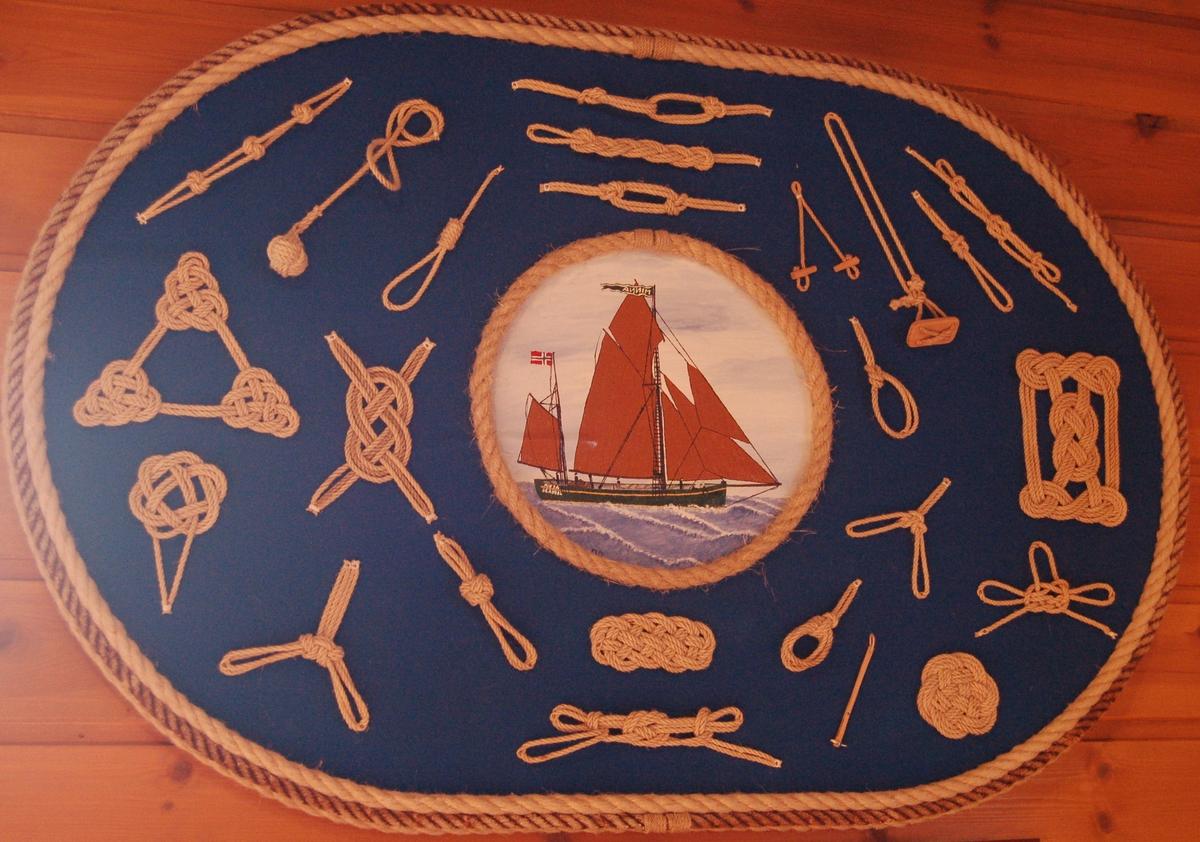 Tavlen er en oval plate med forskellige tauknuter montert på en blå bakgrunn. I midten er der eit
sirkelforma maleri av seglskuta "Minna" innramet av manillatau.
