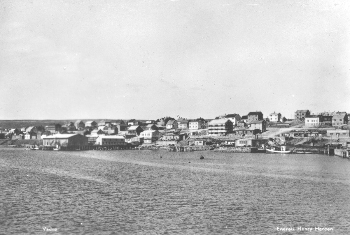 Postkort fra Vadsø. Fra gjenoppbygningen av byen.  Utsikt fra sjøen mot havna, og deler av midtbyen. Det ligger to fiskebåter ved en av kaiene