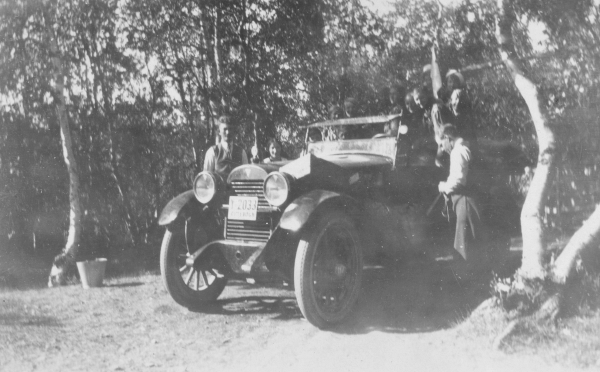 Speidere på hjemtur etter patruljeleir i Rustefjelbma, 4.-10. juli 1930. Bilen de kjørte hadde skiltnummer Y 2033. På bildet ser man to menn som står ved bilen, og oppe i bilen er det speiderjenter. Det er trær omkring. En bøtte står på bakken