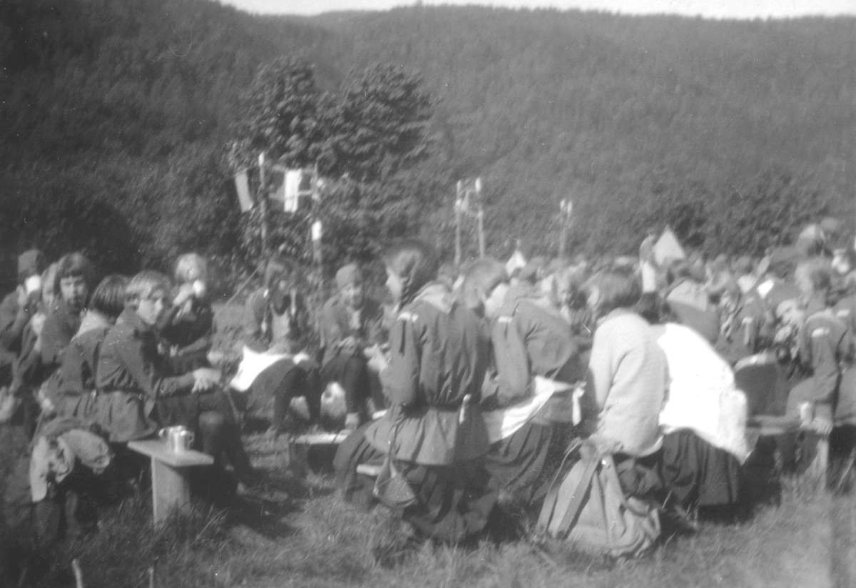 Speiderjenter på pinseleiren i Drammen, 1930. På bildet sitter de på benker. De er kledt i speideruniformer. Det vokser tett skog på fjellet i bakgrunnen