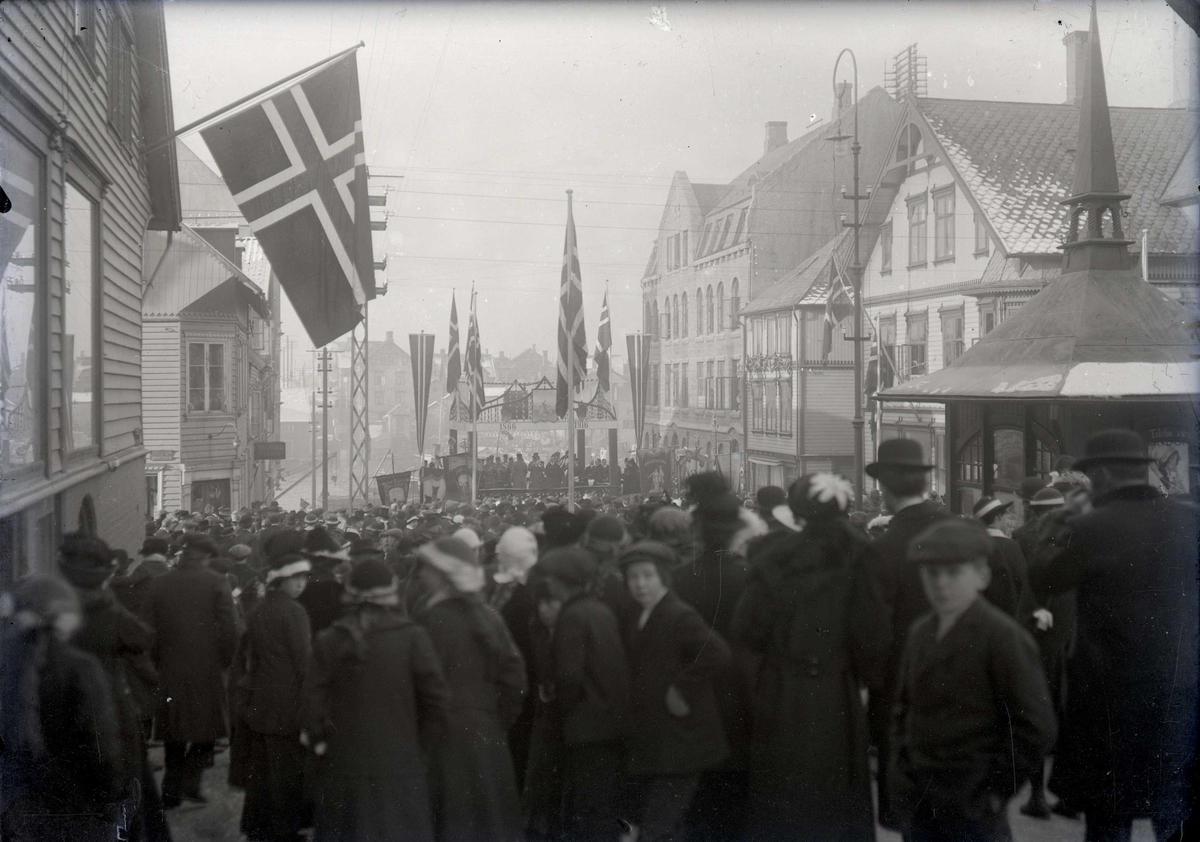 Stor folkemengde samlet i Torggt. Midt i gata bygget opp tribune der det står 1866-1916. Flaggene heist. Feiring av 50-årsjubileet for Haugesund som kjøpstad.