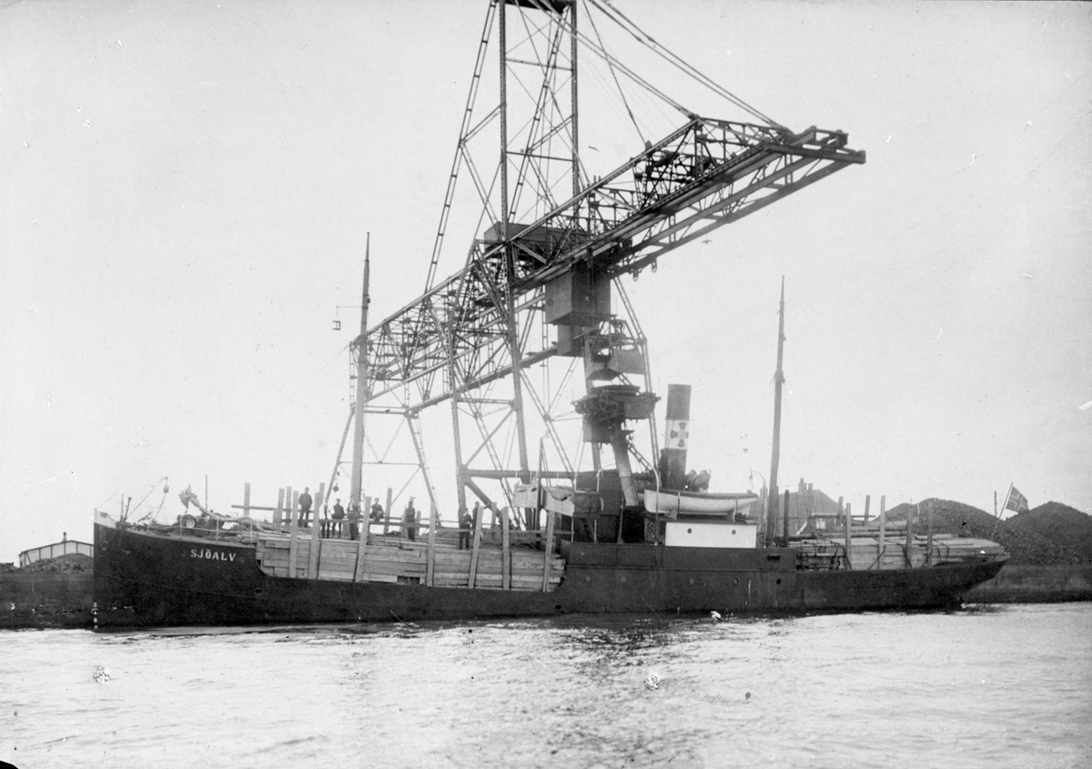 Dampskipet D/S "Sjöalv" under lasting av trelast ved en industrikai. En stor kran står på enden av kaien. Flere mann er på dekk.
