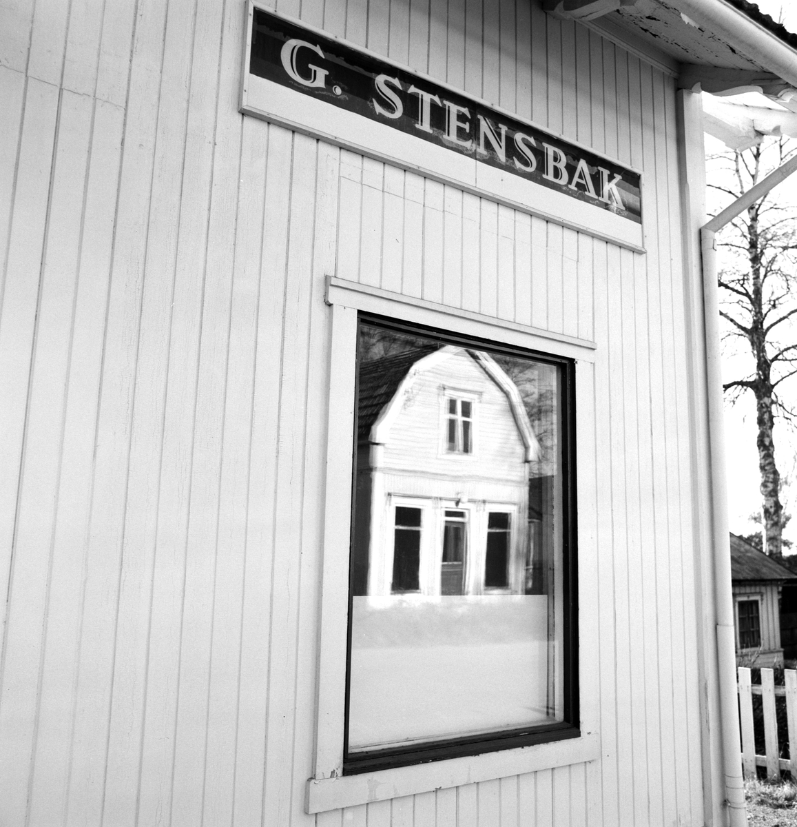 Butikkvindu, skilt med G. Stensbak(slakteforretning og pølsemaker). Klevbakken 83, Ådalsbruk. Bygningen som rommet Jonette Pabsdorfbutikken speiler seg i vinduet. Løten.