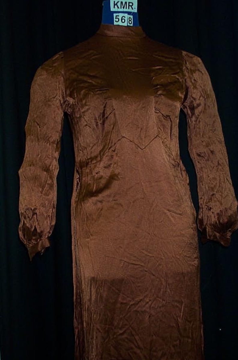 Langermet kjole i gyllenbrun fløyel. Kjolen har høy hals og bånd i sidene til å knyttes bak.