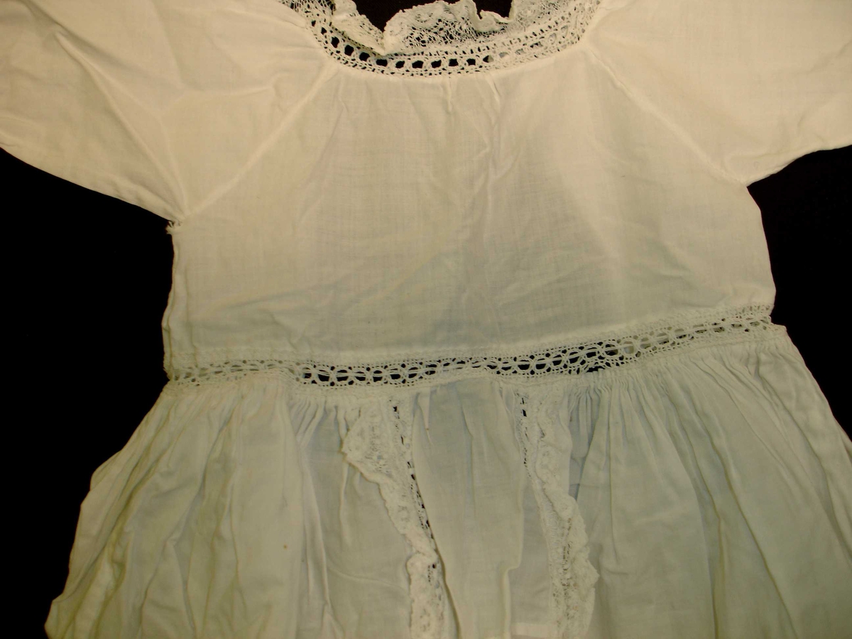 Dukkeklær, kjole i hvit bomullstoff,  blonderkant rundt hals, langs front fra midjen og ned, og ermeåpning. 