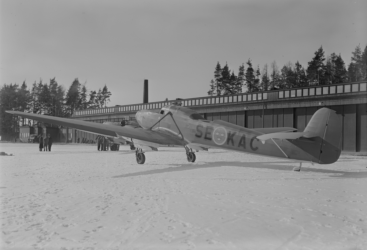 Kartverkets flygplan typ P 6 med registreringsbeteckning SE-KAC, vintertid. Flygplanet sett snett från sidan. Byggnad i bakgrunden.