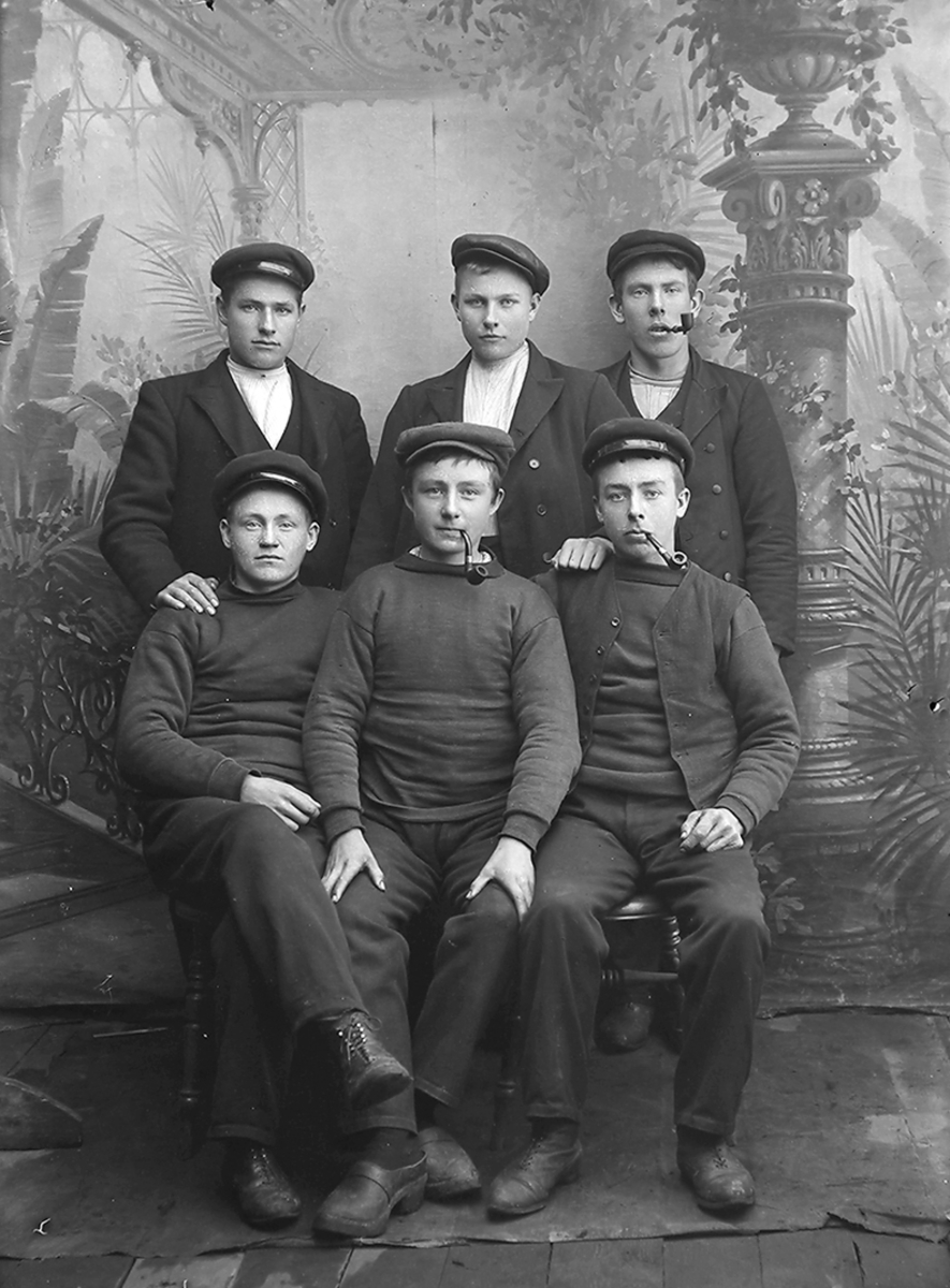 Gruppebilde. Bildet er tatt i Lofoten.Seks menn. Tre sitter, tre står bak. Tre menn har en pipe i munnen. Mennene har på seg genser eller jakke og en lue på hodet.