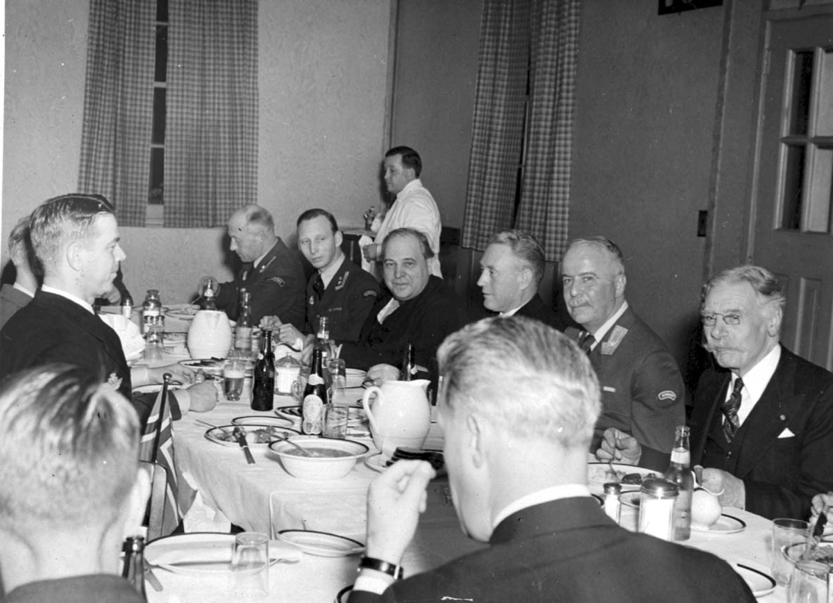 Gruppebilde. Flere personer, menn, de fleste i militæruniform. Sitter ved dekket bord.
