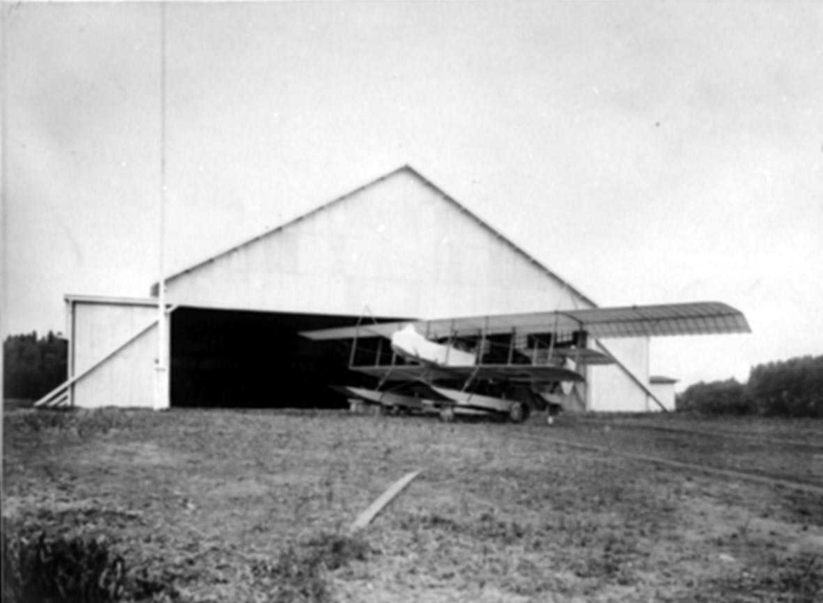 Lufthavn. Ett fly, S.W. Farman med flotører ved en hangarbygning.