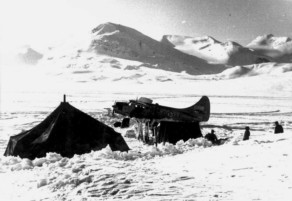 Snødekt landskap. Ett fly på bakken, Otter OAF. To telt, noe utstyr og noen personer ved flyet. 