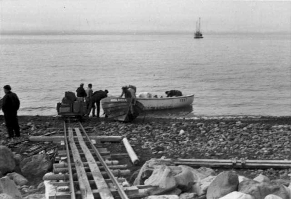 To åpne båter og flere personer ved strandkanten. Jernbanespor i forgrunnen. Fartøy, polarskute i bakgrunnen