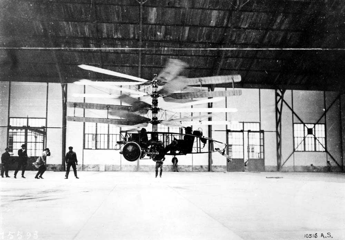 1 helikopter i luften inne i en hangar. Flere personer i hangaren.