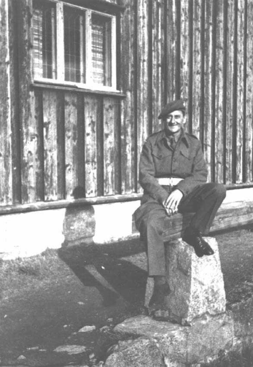 En person. Mann i militæruniform. Sitter på en stein foran en bygning.