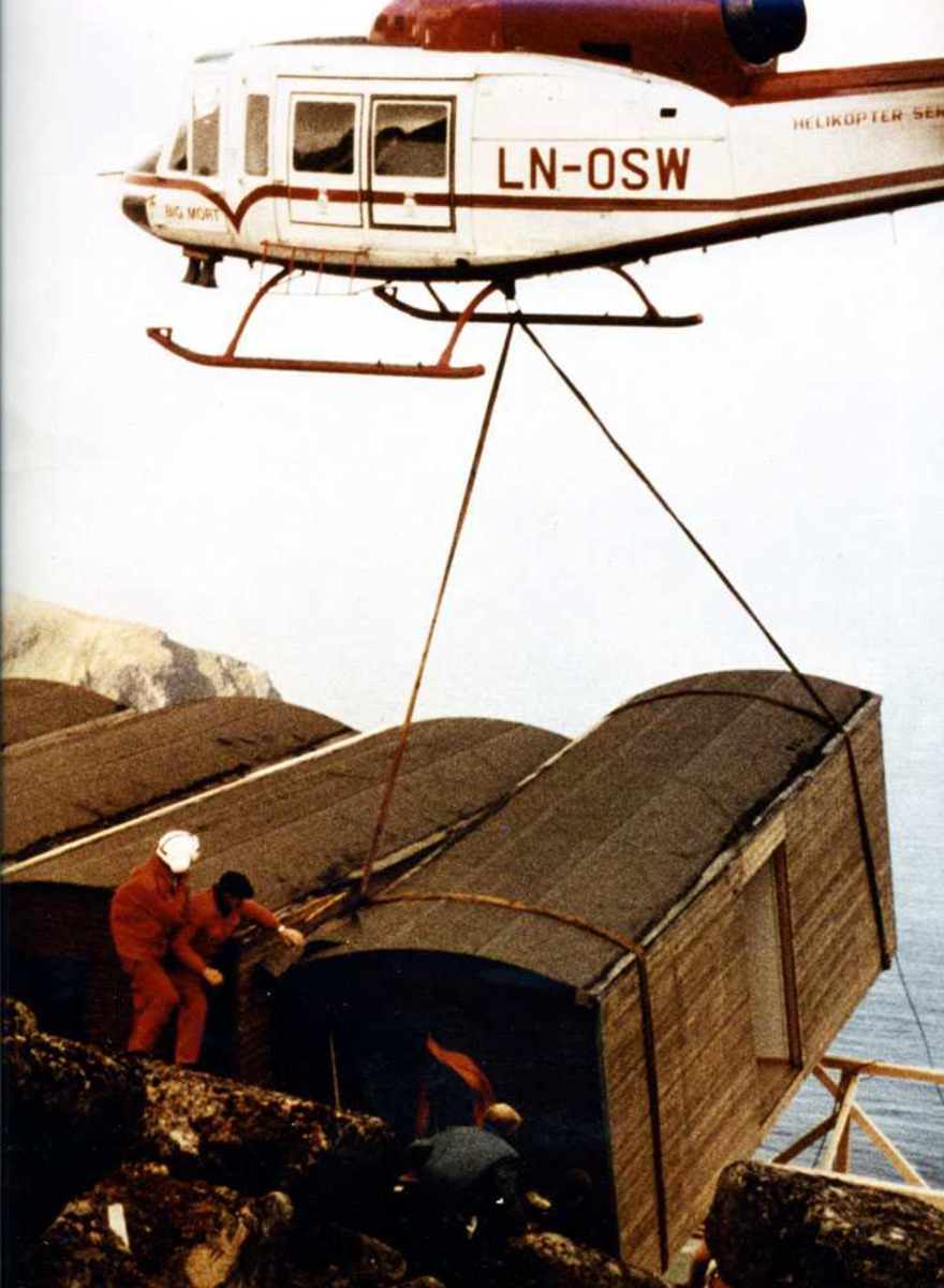 Ett helikopter i luften som løfter/plasserer en anleggsbrakke, LN-OSW. Tre personer ved  brakken. Flere brakker som står ved siden av.