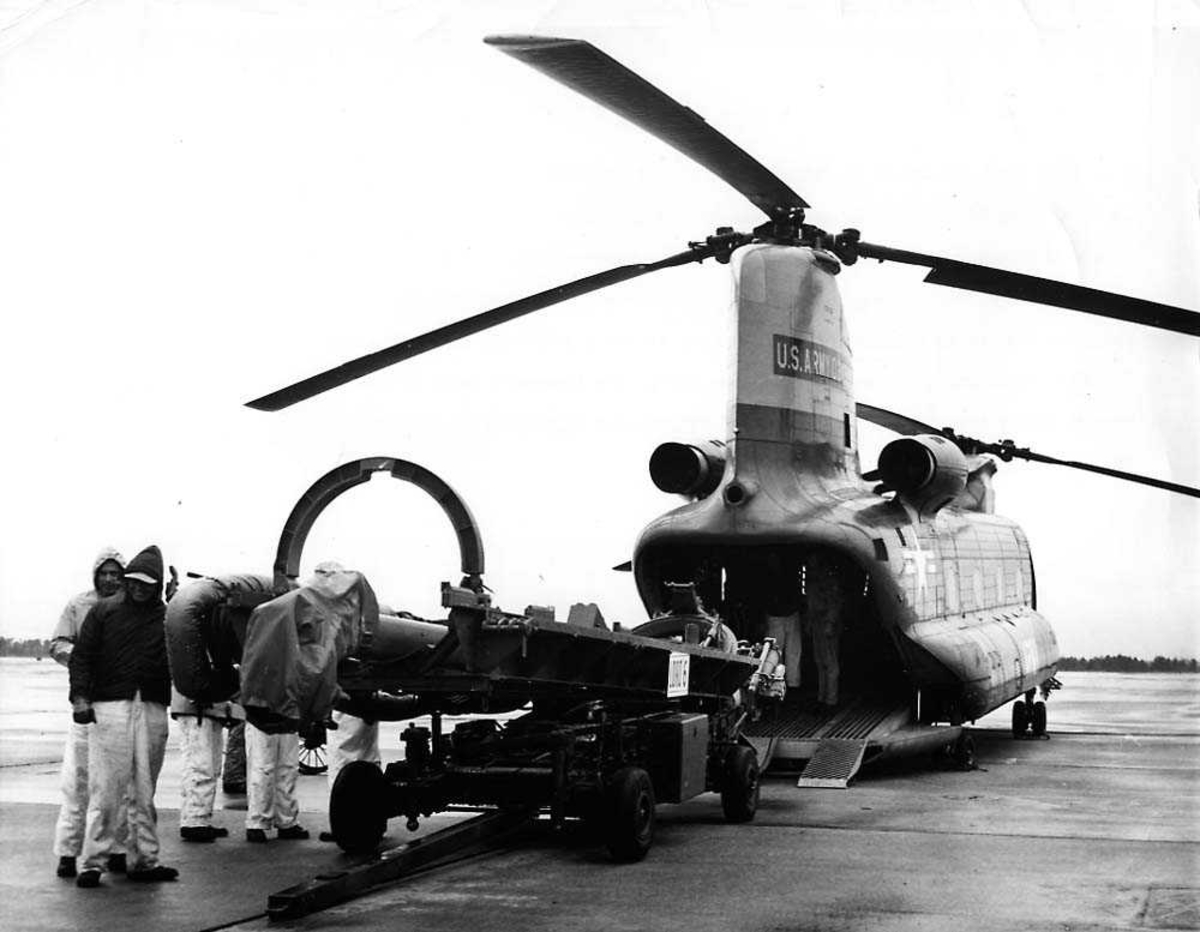 Ett helikopter på bakken, Boeing Vertol Ch-47 Chinook. En utskytningsrampe for missiler ved lasteinngangen til helikoptret. Flere personer.
