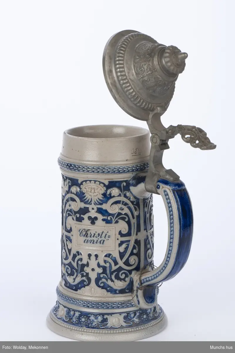 Drikkekrus av Hollands type. Blågrått stentøy med blå dekor. 
Barokkinspirert dekor.
Lokk i metall.