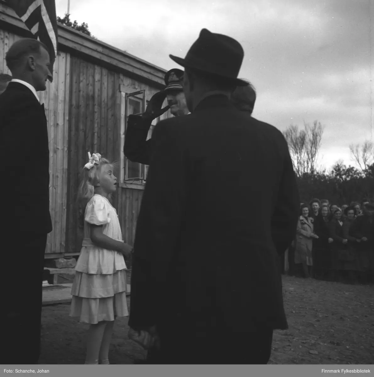 Kongebesøk i 1946:  Kong Haakon VII besøker Tana. Kongen hilser en mann som står halveis med ryggen mot kamera. Foran Kongen står en annen mann slik att vi ser bare ansiktet og hander til Kongen på bildet.  Mellom mennene står en liten jente på hvit kjole, hun har antagelig gitt blomsterbuketten til Kongen.
