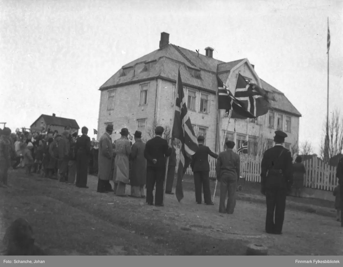 17.mai feires i Vadsø -46. Folk har samlet seg utenfor kysthospitalet / sykehuset  med paroler og flagg. (alle står med ryggen til mot fotografen).  Lengst nede i høyre hjørne sitter en hund (bare delvis med på bildet).