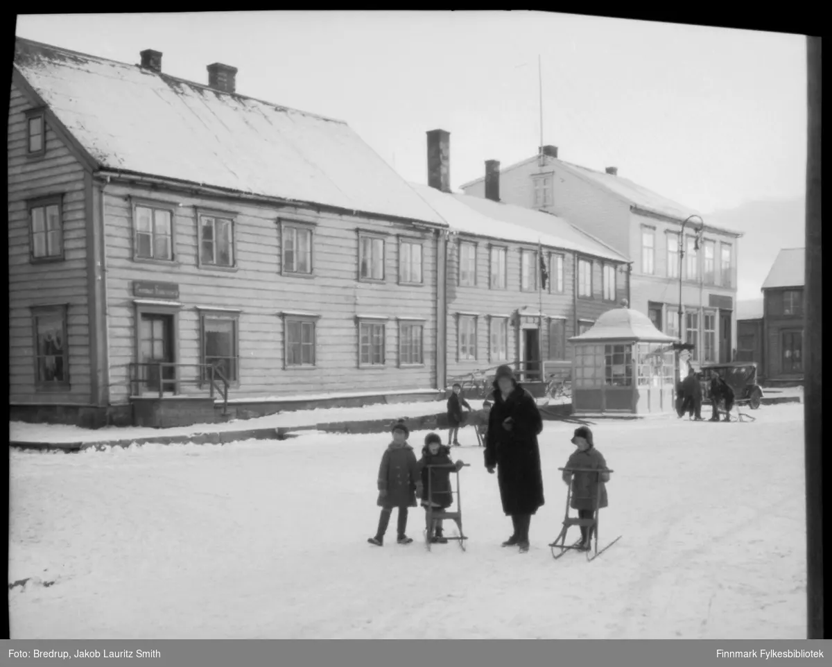 Vinterbilde fra torget i Vadsø.  Antakelig er kvinnen på bildet Hildur Bredrup, og noen av barna er Bredrup-barn.  De bruker spark.  I nærheten av kiosken en liten gruppe gutter og menn, og en bil står parkert.  Butikker og forretninger ved torget.