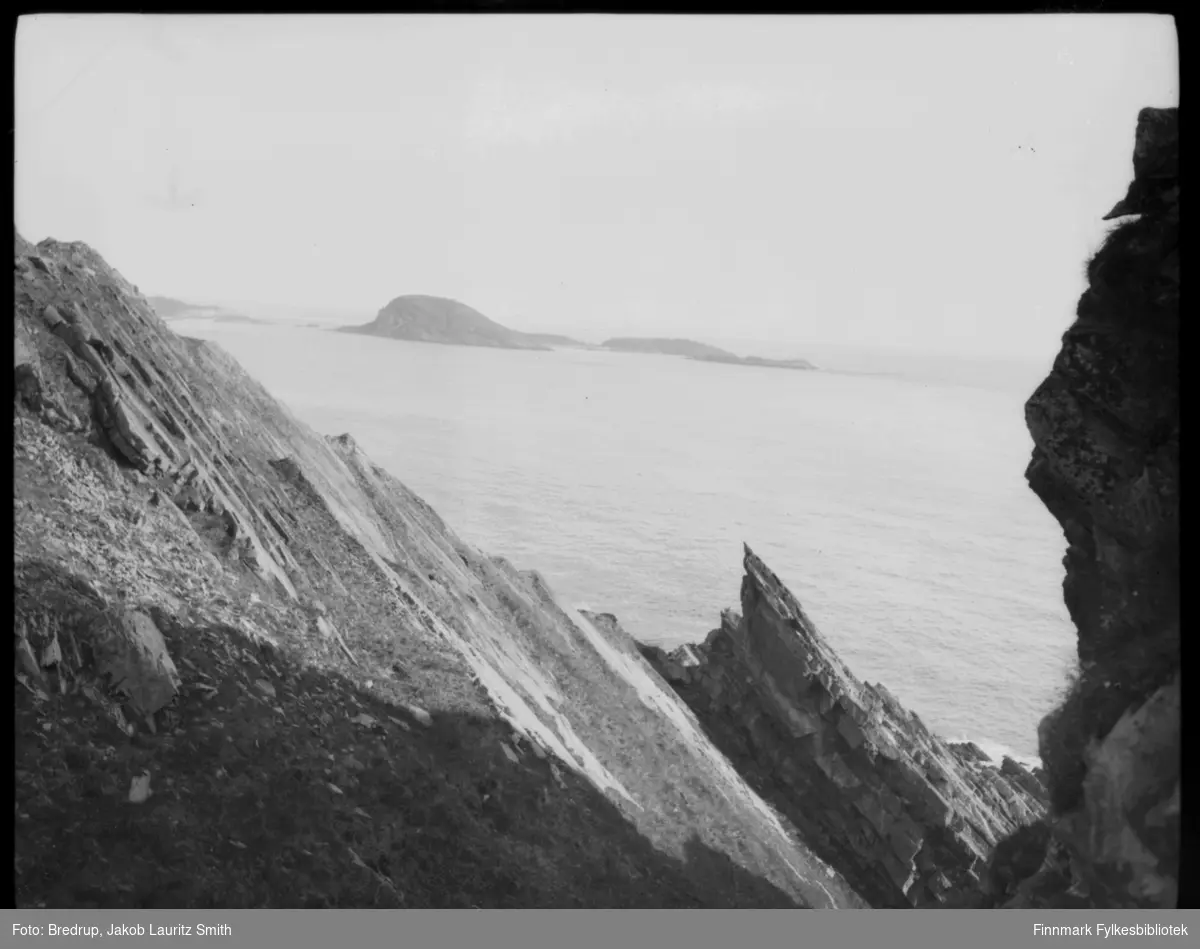 Fotografen har stått mellom noen klipper og tatt et oversiktsbilde utover noe som kan være Kongsfjord med Veidnesodden og øyene Kongsøya, Skarvholmen og Helløya.