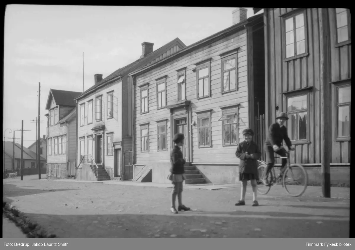To jenter leker i Theatergaten i Vadsø og en mann sykler forbi.  Huset nederst i gata jentene leker i, er Kildahl Olsens bokhandel.  Lengst borte fra kamera møter Theatergaten Havnegata.
