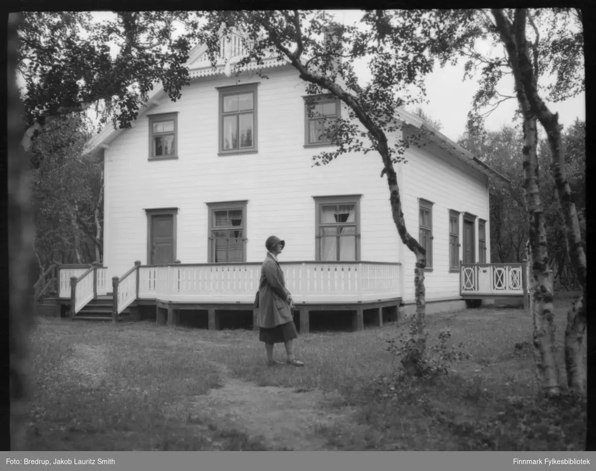 Hildur Bredrup fotografert foran et stort, lyst hus, omgitt av høye trær.  Hun er kledd i drakt og hatt.  Er huset det samme som på Fb.98012-213?  Huset er omgitt av en stor veranda.
