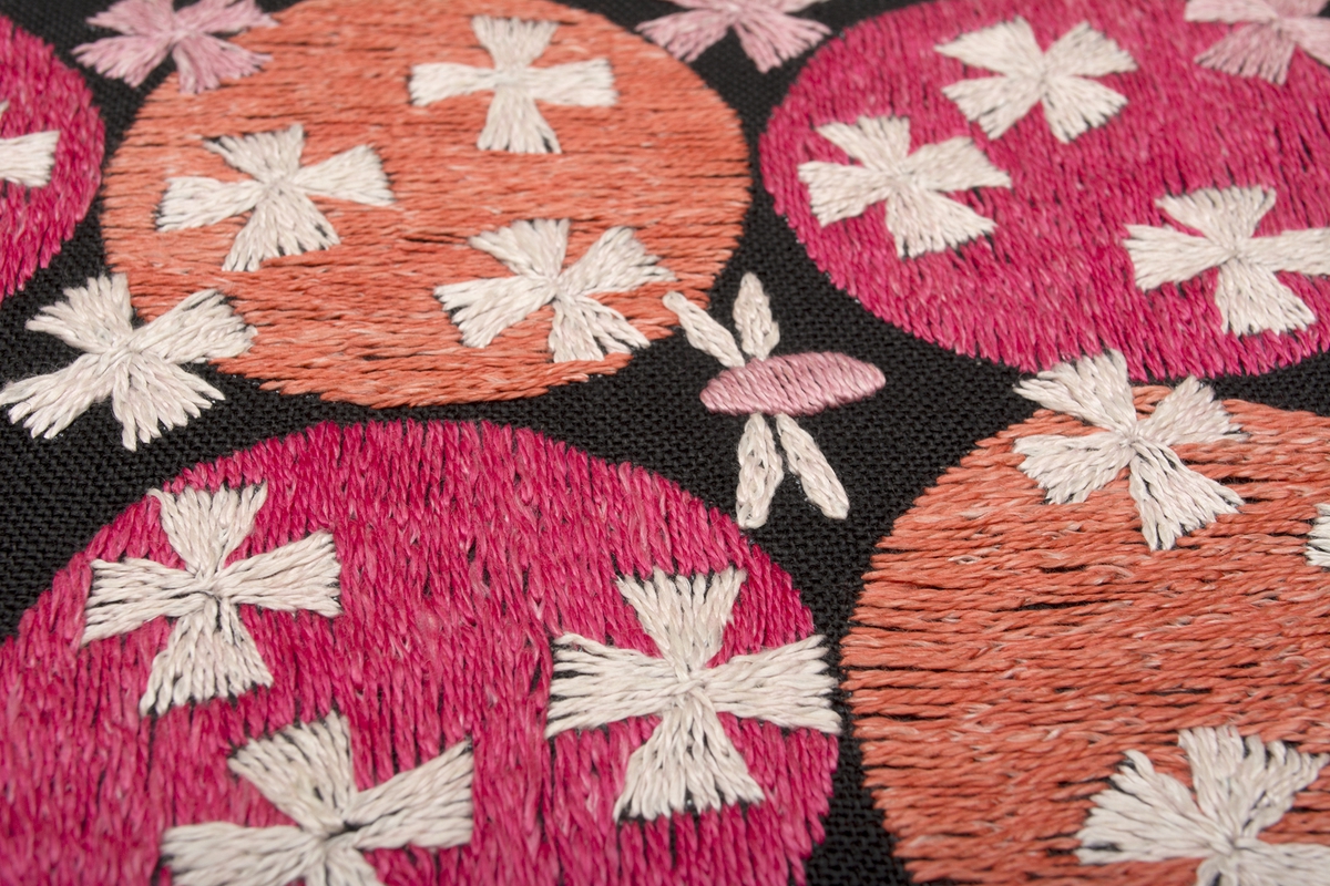 Rektangulär duk på svart linnebotten, fodrad med blekt linnetyg. Motiv med tre x fyra cirklar (en cirkel = 67 mm) i två olika rosa lingarner. Två flygfän fladdrar omkring. Fyrbladiga blommor över både rosa cirklar och svart botten.