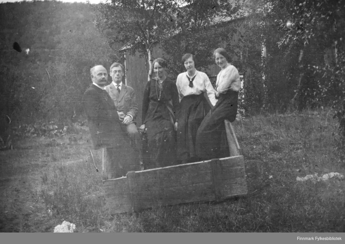 Fem personer, tre damer og to menn, fotografert mens de sitter på kanten av en trekasse i Rassejåkka i Tana. Alle sammen har på seg fine klær. I bakgrunnen ser man en bygning mellom trærne.