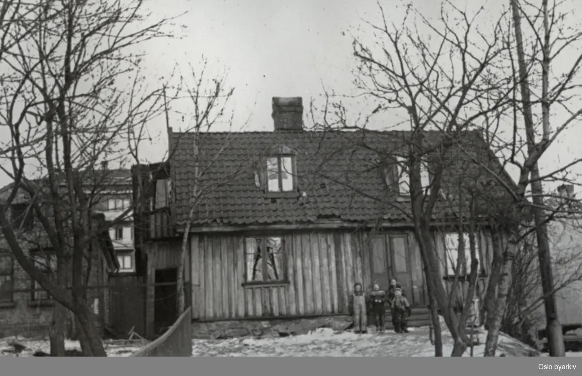 St. Halvardsgate 34. Fire vinterkledde gutter står ved inngangsdøren til det lille trehuset. Fra fotoalbum med tittel; Oslo kommune leiegårder, nedrevne eiendommer.