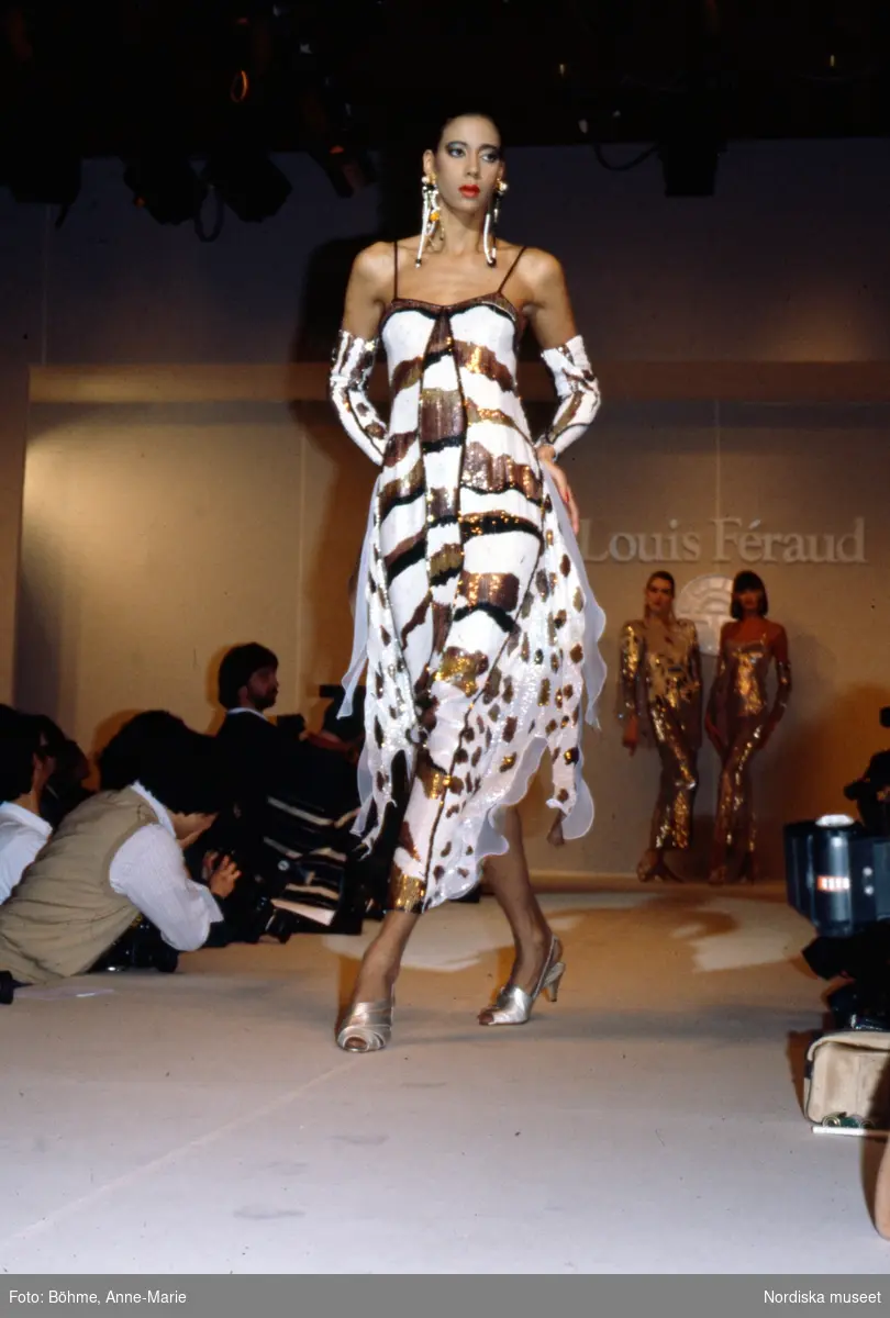 Modevisning. Modell i vit klänning med svart- och guldfärgat mönster, sandaletter i silver och matchande örhängen. Från Louis Féraud.