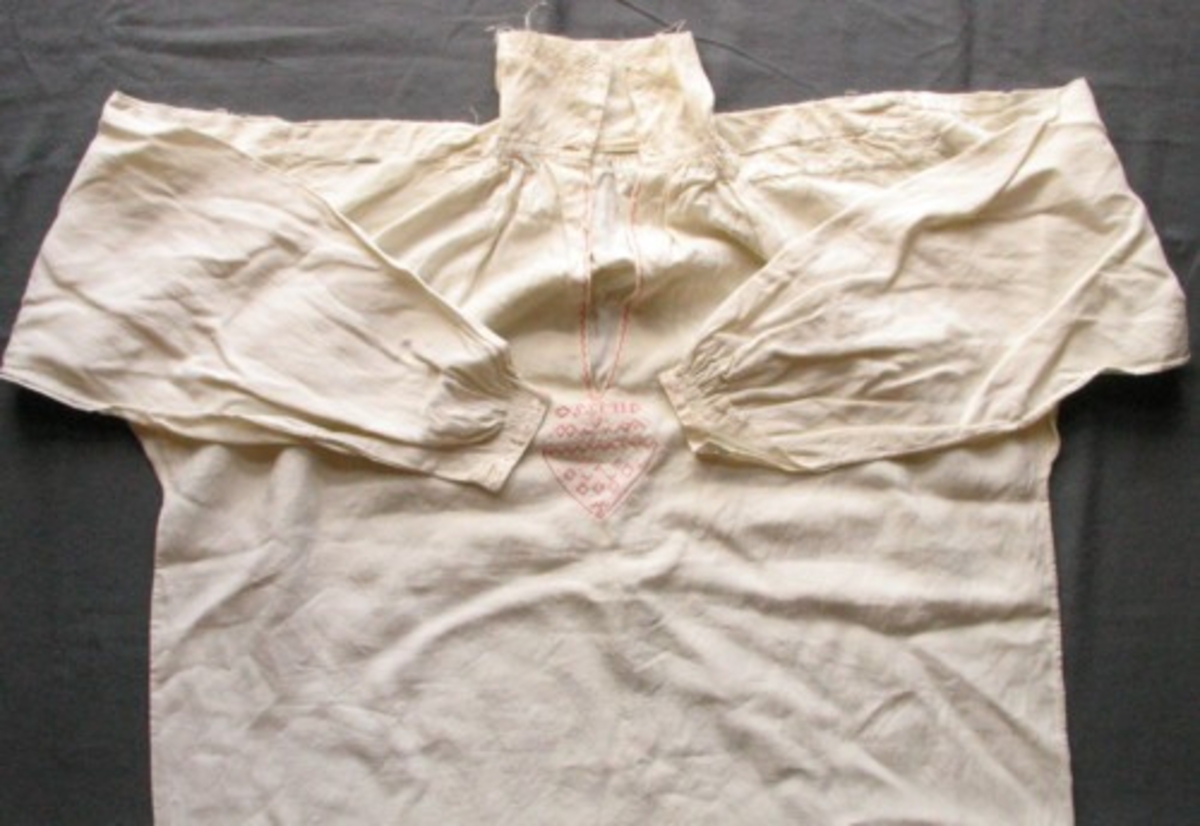 Äldre mansskjorta i handvävt linne, förmodligen brudgumsskjorta med broderat hjärta (80 X 85 mm, i korsstygn och stjälkstygn) under sprundet framtill (215 mm djupt, kantat med rosa broderier) och med märkningen "OSSMID" (9 X 62 mm) i nu rosa bomullsgarn. Krage med broderi i vitt merceriserat bomullsgarn (90 mm), broderi som bård längs kragens kanter i rätlinjig plattsöm som sträcker sig ca 20 mm in på kragen. Förstärkt ok, broderi på oket från hals till axel, rynk fram och bak vid kragen, broderi för att förstärka rynktråden. Broderi vid ärmisättningen, ärmspjäll. Stripade rynkor vid manschetten (19 mm bred), broderi på manschetten både i vitt merceriserat bomullsgarn och rött bomullsgarn, två knapphål, förstärkta ärmsprund (tyget invikt i en trekantsform). Sprund nertill i sidsömmarna (86 mm).