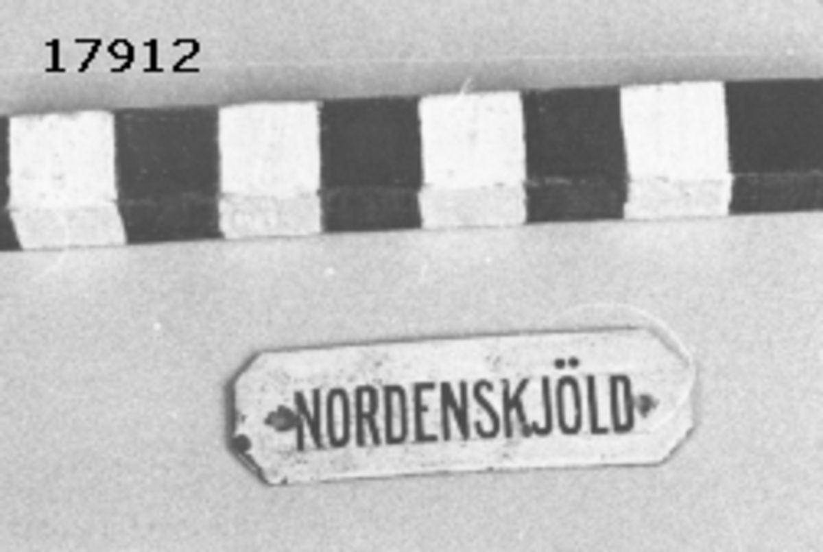 Rekangulär skylt av plåt med avklippta hörn. Framsidan vitmålad och försedd med handmålad text i svart: " NORDENSKJÖLD ". Hål i kanterna för spikar.