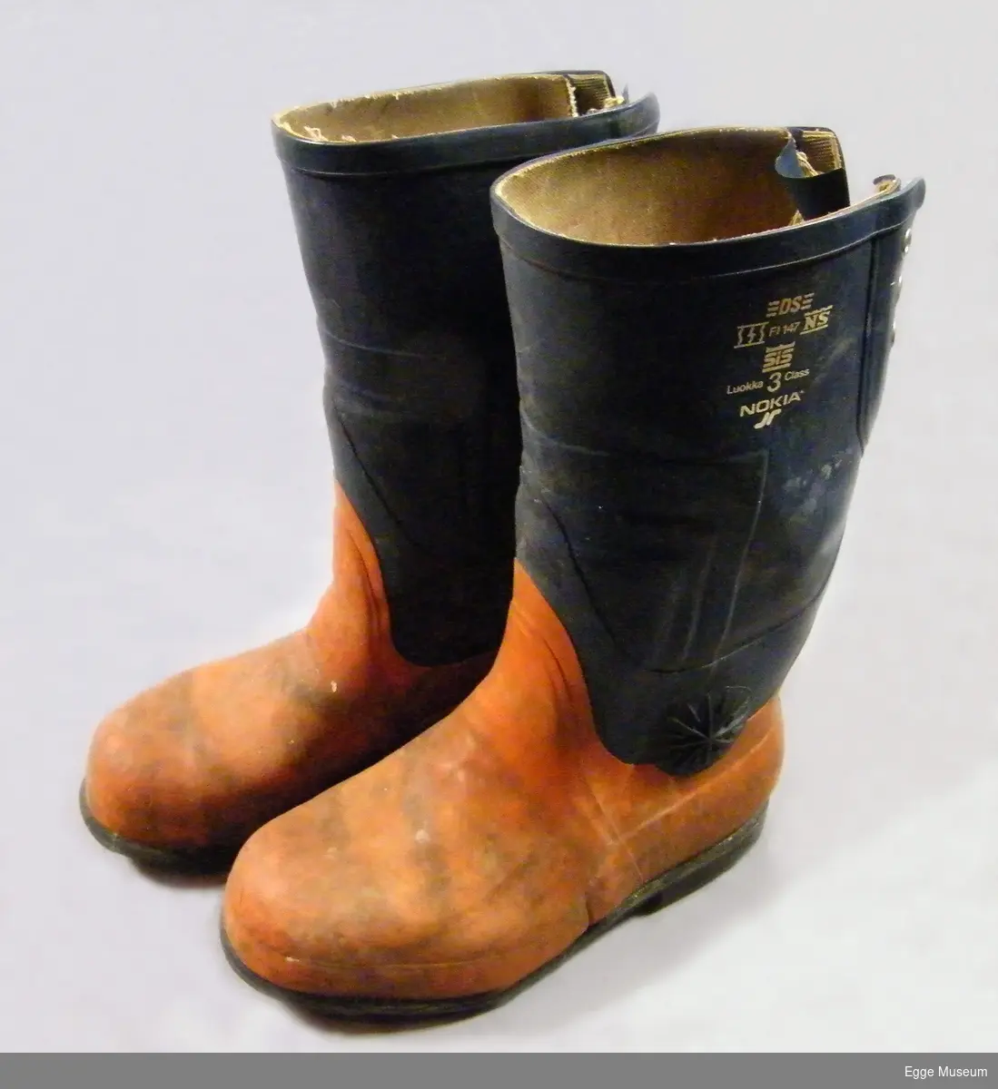 Vernestøvler for skogsarbeid fra 1980-tallet. Utvendig er støvlene trekt med gummi. Gummien er limt og sveiset sammen, muligens smeltet sammen over de områdene hvor gummien er tykkest. Flere lag? Tykkest gummi over typiske viktige verneområder, som tå- og hæl. Det er også innstøpt et stålparti i tåpartiet. Noe tykkere også oppover deler av legg og vrist. Anklene er beskyttet med pålimte gummiringer (i stjernemønster). Begge støvlene har skader av motorsag. Skaftet i støvlene er blått, foten i oransje farge. Støvlene kan snøres sammen med lisser på baksiden av støvlene (toppen av skaftet). Kraftig gripemønster på sålene med små hull (beregnet for pigging av støvlene). Foret med lerretsstoff. Ingen løse såler. Størrelse 41.