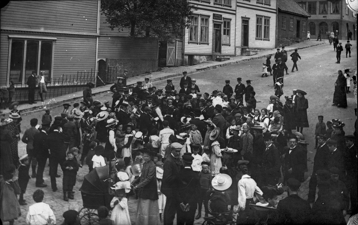 Mange personer på gaten nede ved Torvbakken, bl. a. flere barn. Et orkester som spiller midt på bildet. Bolighus og fortau  i bakgrunnen.