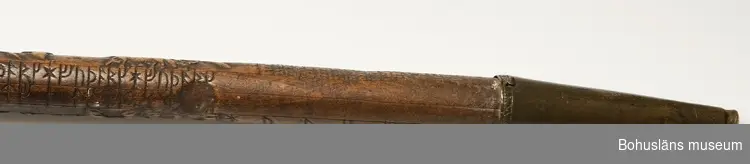 Käpp tillverkad av en runstav, kompletterad med krycka av elfenben samt doppsko av mässing. Skador på kryckan; sprickor samt del av handtaget bortslaget.
Runstaven är troligen tillverkad av björk, skurna figurer i högrelief, runor skurna i lågrelief; arbetet svärtat och polerat.
Runstaven daterad till 1600-talet.

Tullkontrollör A. Kidron var sedermera tullförvaltare i Kalmar.
Möjligen har tullkontrollör Kidron fått fel begynnelsebokstav på förnamnet. I Sveriges statskalender/1881 finns en Kidron antecknad i Kalmar: Tullförvaltare, Carl Erik Reinhold Kidron, R. W. O., f. 26; 79; utöfvar jemväl chefskap öfver kustbevakn. i Kalmar tullkammar distr.  

Se artikel av Sten Kristiansson i klipp-pärmen Glimtar från Uddevalla museum med artiklar om Uddevalla museum publicerade i Bohus-Posten åren 1940-1942; Runstavar.
Bohusläns museums bibliotek, NcbkzN-Uddevalla

I 1869 års tryckta katalog G N:o 20:
En runstaf med elfenbernskrycka och upphöjda figurer.

Ur Nationalencyklopedin, NE.se: Runkalender
Runkalender, en form av evighetskalender (jfr kalender), där beteckningarna för veckodagarna och gyllentalen utgörs av runor: futharkens sju första runor (upprepade 52 gånger) resp. futharkens 16 runor utökade med tre nyskapade tecken (placerade vid de datum då nymåne beräknas inträffa under vederbörande år i den 19-åriga måncykeln). Även de kyrkliga festdagarna kan vara angivna med runor. Runkalendrar har förekommit i olika utföranden, bl.a. skrivna på pergament eller ristade på trä, ben eller horn. Runkalendern tycks vara en medeltida svensk uppfinning, medan träkalendrar utan runor, men med dagarna markerade med streck eller hack i stavens kant, är kända från flera områden i Europa. Den äldsta bevarade runkalendern, ristad på en trästav, har daterats till 1200-talet (Nyköpingsstaven). Det tusental kalendrar av trä som är kända tillhör företrädesvis 1500- och 1600-talen. Under 1700-talet fick runkalendrar en renässans, och omkring 1800 blev det populärt med liknande kalendrar i form av tobaksdosor av mässing. Runkalendern kallas ibland primstav och runstav.