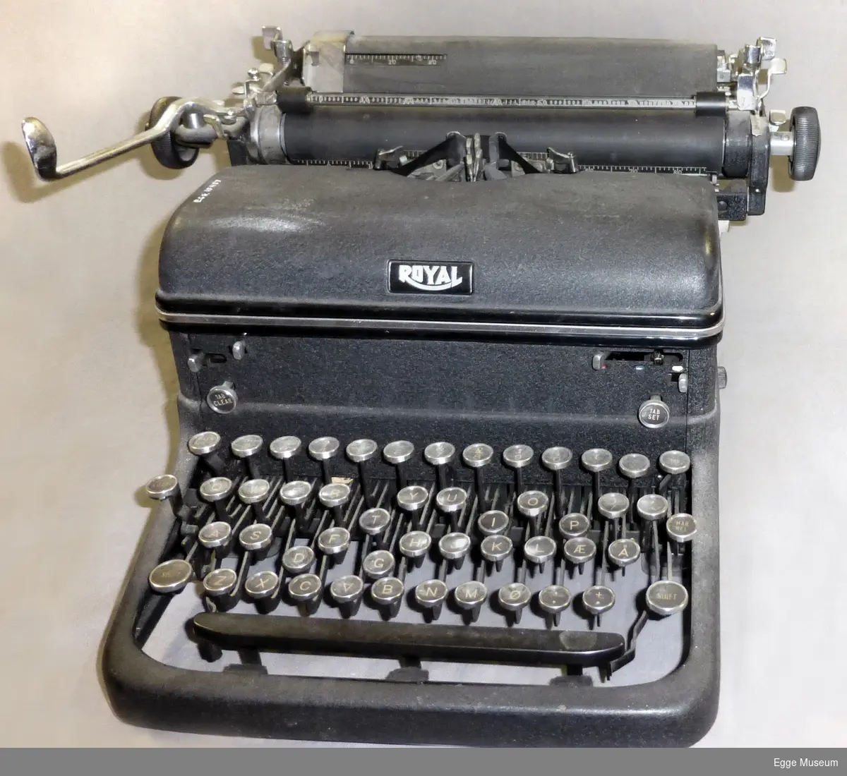 Roayl skrivemaskin, produsert i New York, USA. Metallhus med deksel over stempelarmer. Runde taster. Rustfritt stål i bevegelige knapper og armer. Maskina har innspeksjonsluker på hver side.