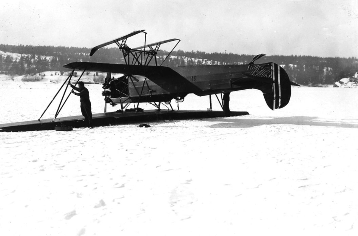 Åpen plass, to personer ved ett fly som ligger på bakken, Fokker CVE nr 311, med skiunderstell. Snø på bakken. Flyet ligger på "rygg".