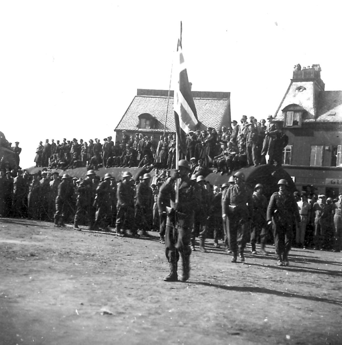 Frigjøringsdagene i Bodø etter krigen 1940 - 1945. Fra kaiområdet, stor sylinderformet "tank" foran noen bygninger. Mange personer samlet på tanken. En gruppe soldater oppstilt foran.