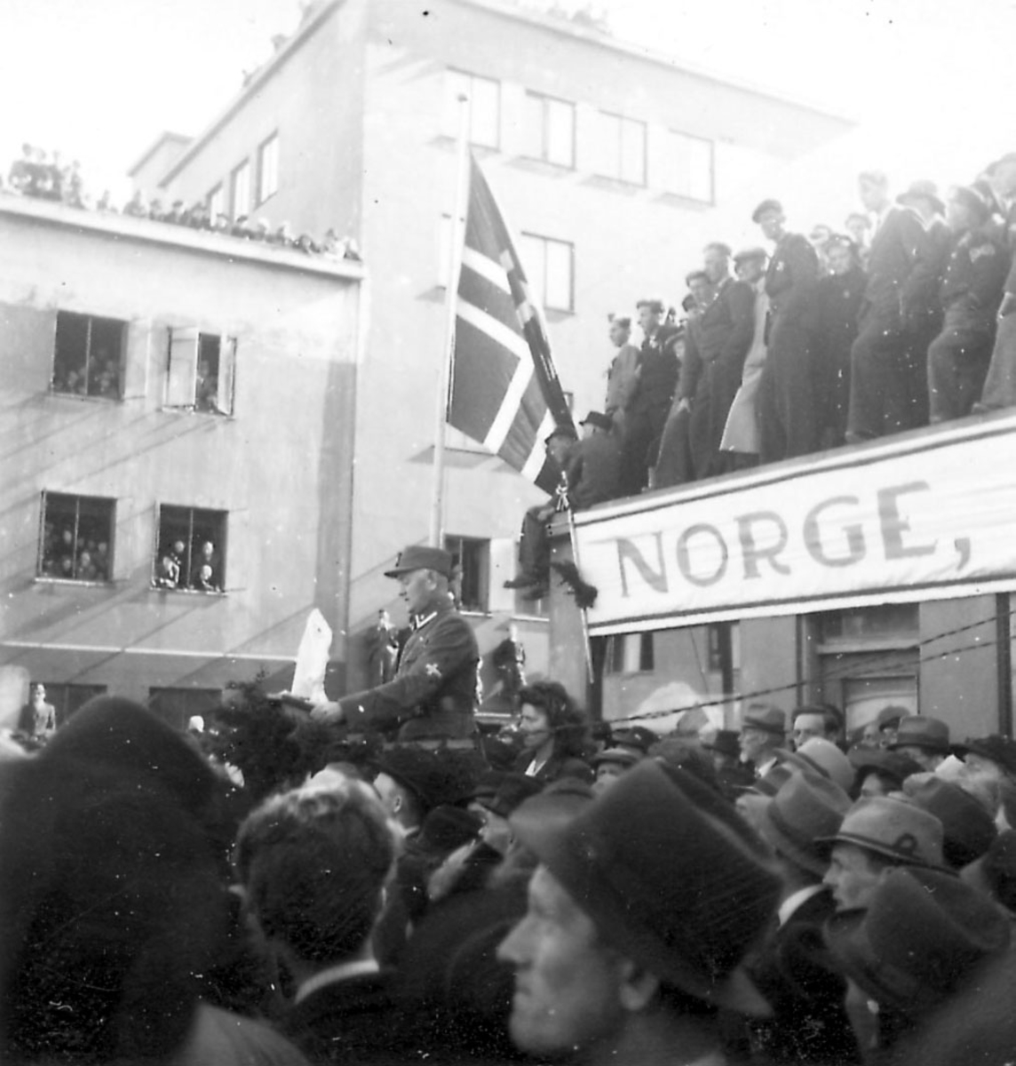 Frigjøringsdagene i Bodø etter krigen 1940 - 1945. En person/offiser holder tale. Mange personer er samlet. Margarinfabrikken i bakgrunnen.