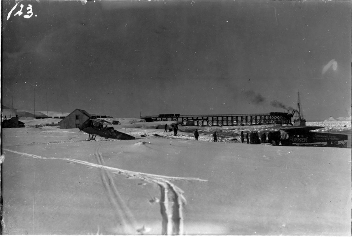 2 fly, T.v. Curtis, T.h., ved strandkanten, Fokker FVII "Fosephine Ford". Flere personer i arbeid. Kai bak med fartøy, KNM "Heimdal". Bygninger bak. Snø på bakken