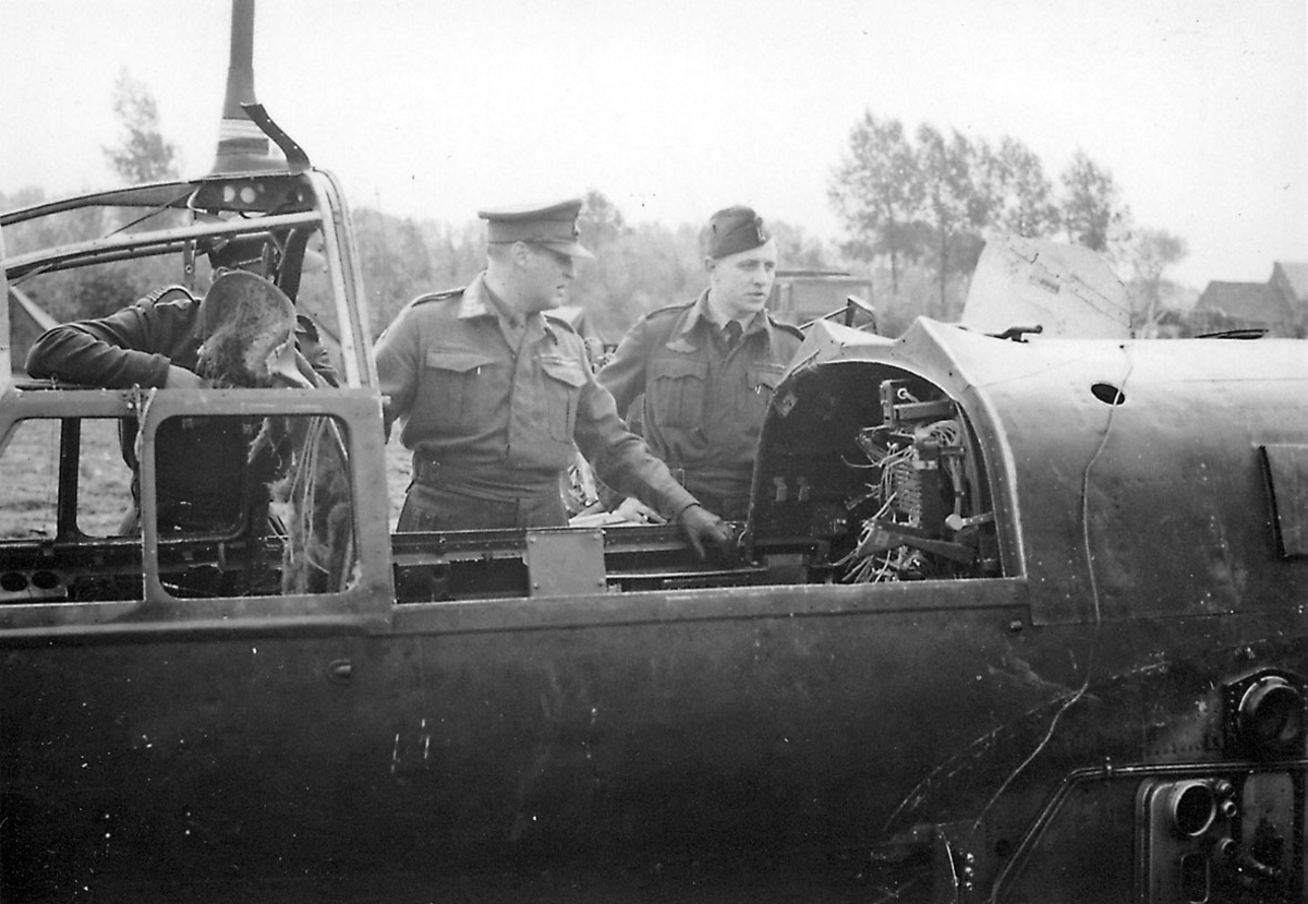 3 personer i militæruniform utendørs, Kronprins Olav i midten. Studerer cockpiten på et fly, sansynlivis et flyvrak.