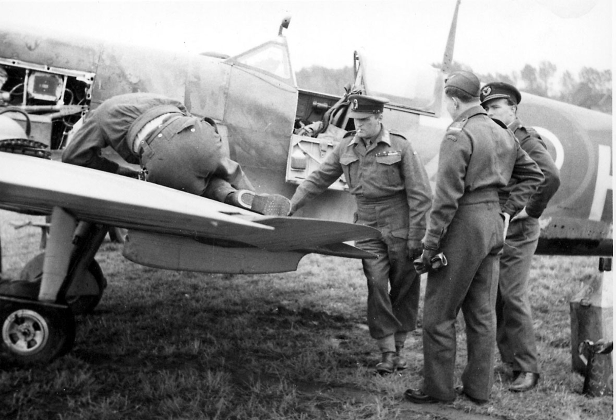 4 personer i militæruniform. Kronprins Olav og 3 andre "studerer" et fly.
Flet en Spitfire