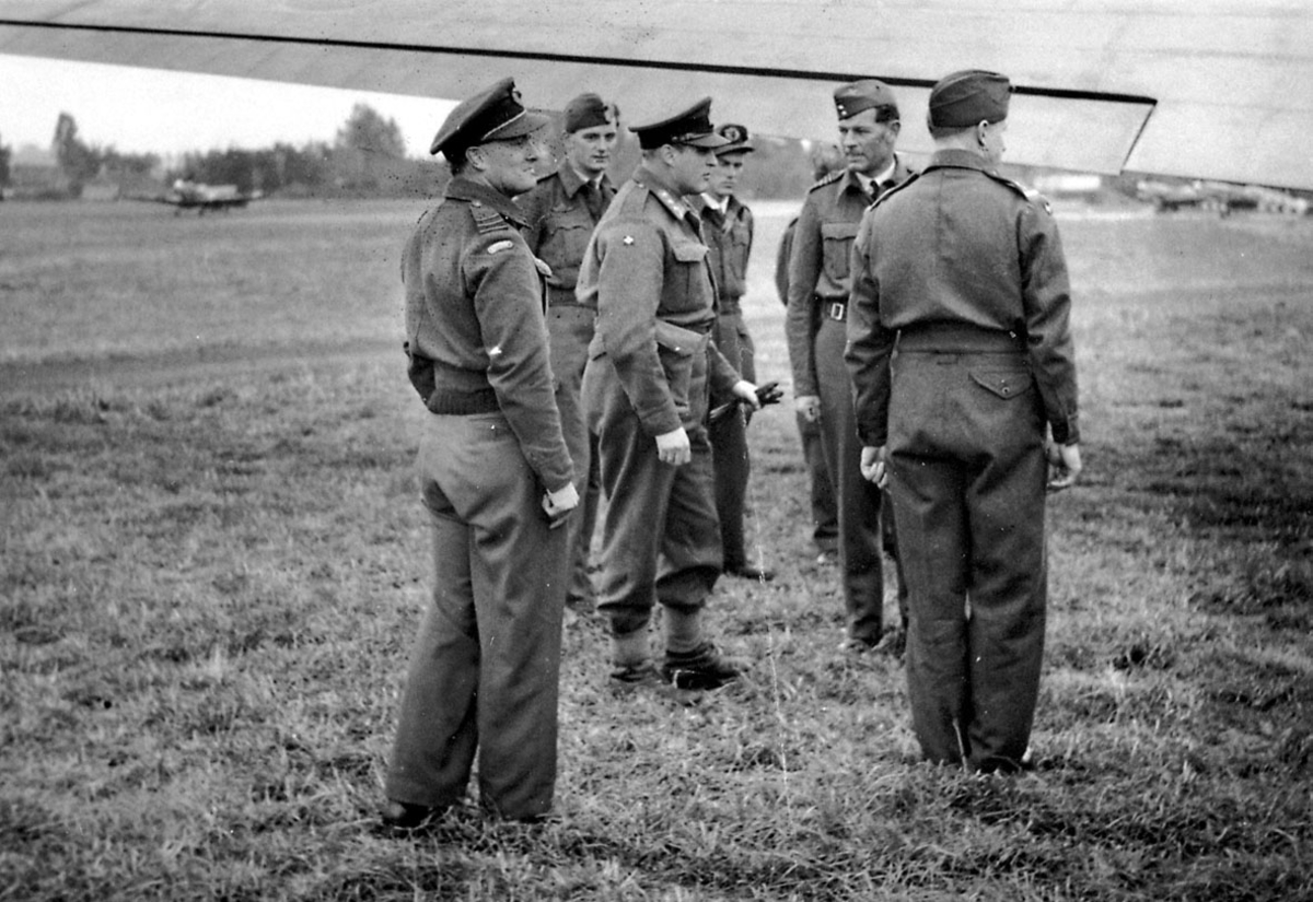 7 personer i militæruniform. Kronprins Olav sammen med 6 andre, på en åpen plass, under en flyvinge. Noen fly i bakgrunnen. 