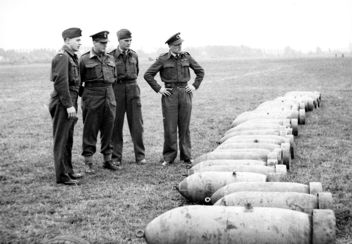 4 personer i militæruniform. Kronprins Olav og 3 andre, på en åpen plass. Inspiserer flere flybomber som ligger på bakken.