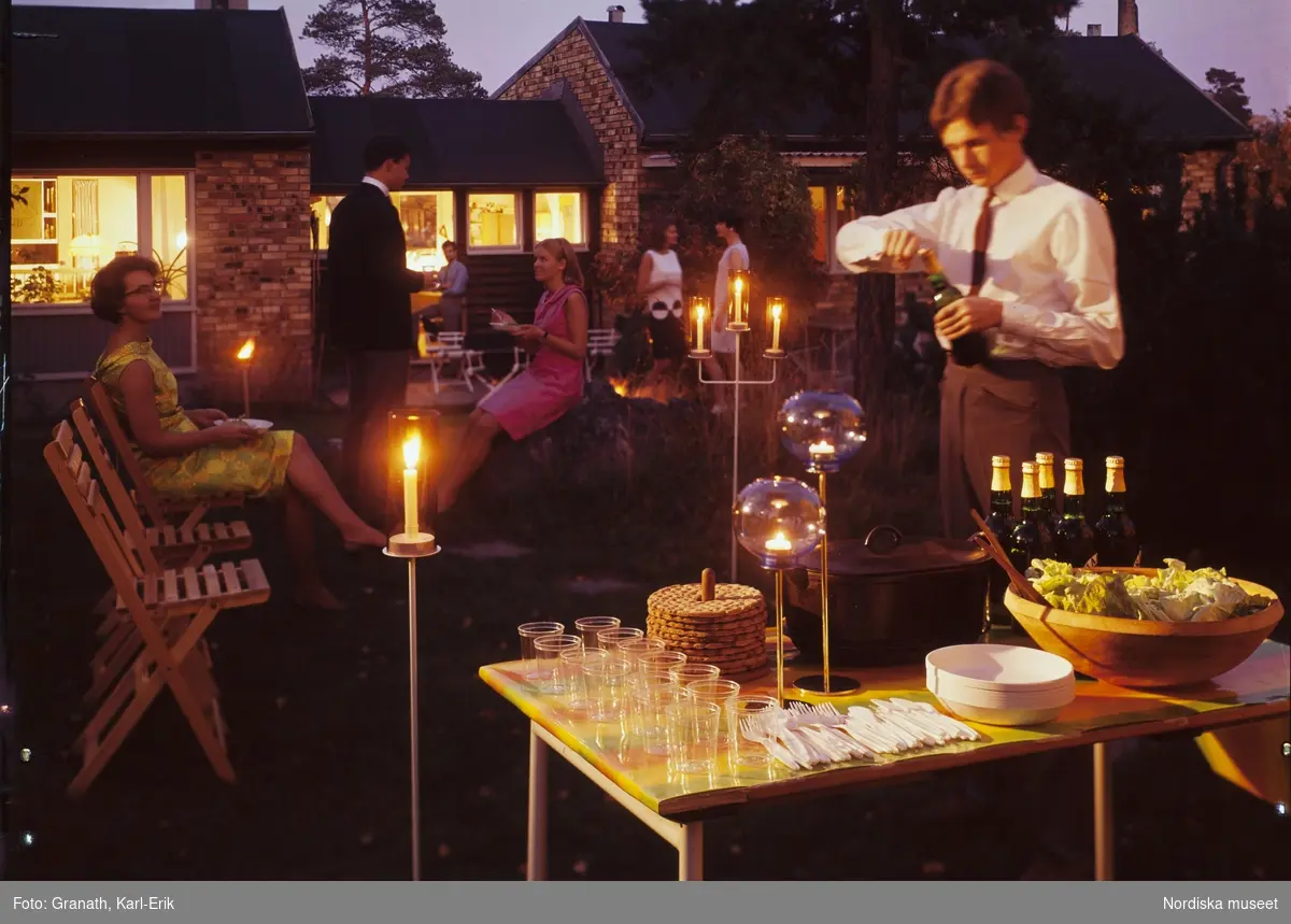 Ett sällskap i en trädgård. I förgrunden ett dukat bord med glas och tända ljus bl a. En man öppnar en ölflaska. I bakgunden sällakap i samtal  framför en upplyst villa i gult tegel.