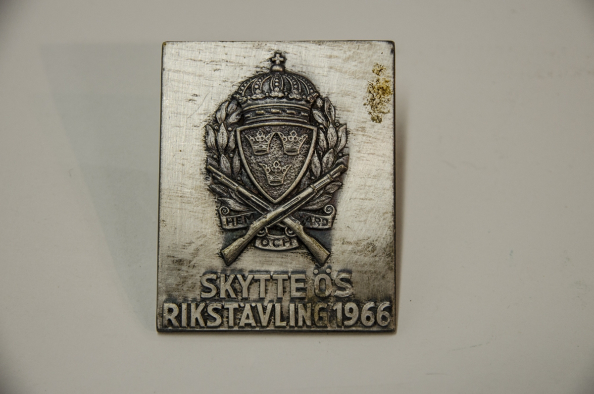 Idrottsplakett i metall med motiv: vapensköld med korsade gevär och texten Hem och Härd. Skytte ÖS Rikstävling 1966.